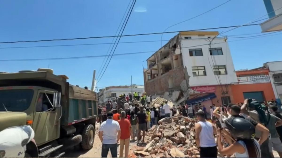 Crowd and rescue workers gather around a collapsed multi-story building with rubble on the street awaiting clearance or rescue efforts.