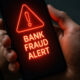 Smartphone screen glows red with a neon warning: 'BANK FRAUD ALERT', held by a hand with a smartwatch visible on the wrist.