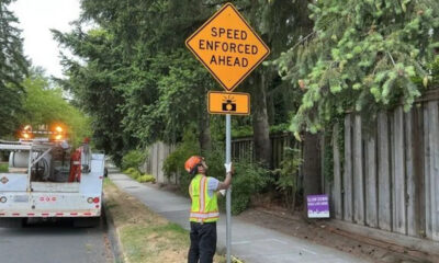 Worker in a hi‑vis vest and orange helmet holds a 'Speed Enforced Ahead' sign beside a sidewalk, with a utility truck in the background.