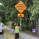 Worker in a hi‑vis vest and orange helmet holds a 'Speed Enforced Ahead' sign beside a sidewalk, with a utility truck in the background.