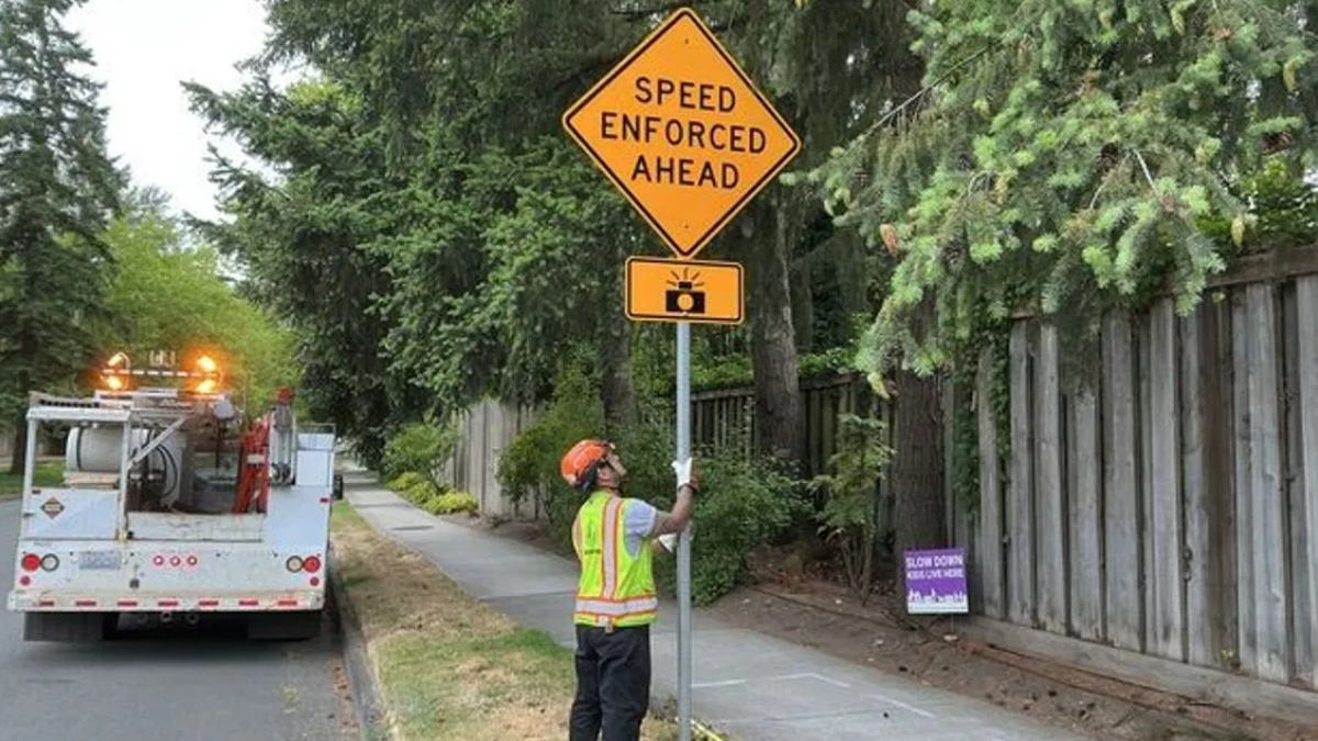 Worker in a hi‑vis vest and orange helmet holds a 'Speed Enforced Ahead' sign beside a sidewalk, with a utility truck in the background.