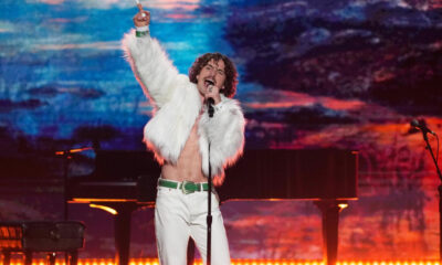 Male singer in a white faux-fur jacket and white pants performs on a brightly lit stage, microphone in hand, raising one finger in the air.