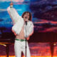 Male singer in a white faux-fur jacket and white pants performs on a brightly lit stage, microphone in hand, raising one finger in the air.