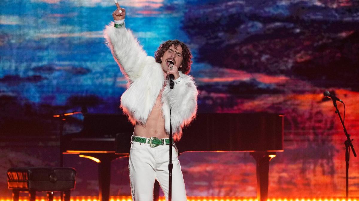 Male singer in a white faux-fur jacket and white pants performs on a brightly lit stage, microphone in hand, raising one finger in the air.