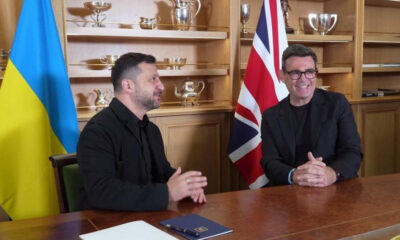 Two men sit at a wooden table in a formal meeting, Ukrainian flag on the left and British flag on the right, trophy-filled shelves behind them.