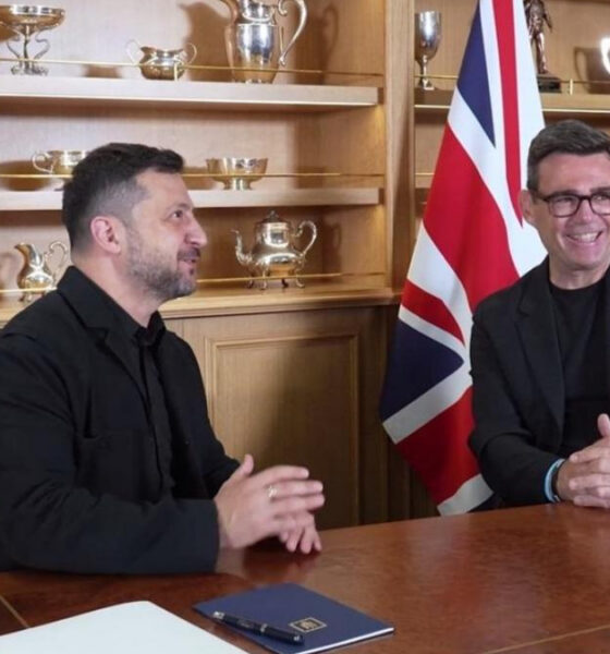 Two men sit at a wooden table in a formal meeting, Ukrainian flag on the left and British flag on the right, trophy-filled shelves behind them.