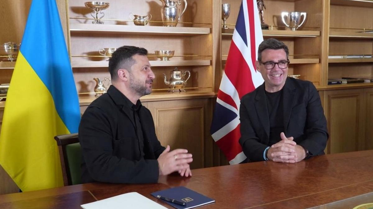 Two men sit at a wooden table in a formal meeting, Ukrainian flag on the left and British flag on the right, trophy-filled shelves behind them.
