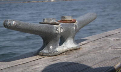 Metal mooring cleat bolted to a weathered wooden dock with water in the background.