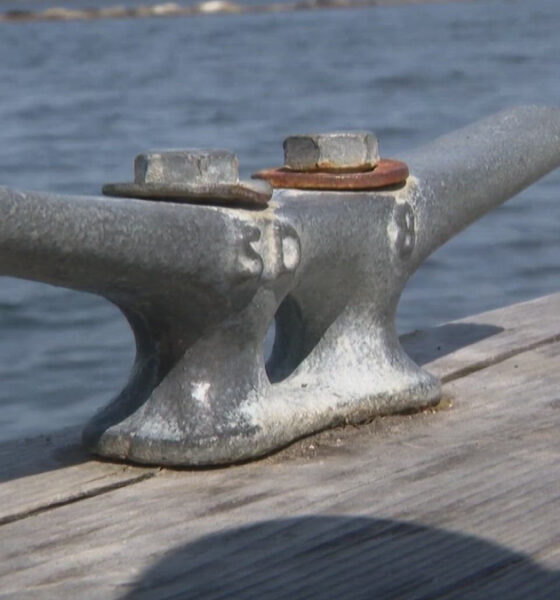 Metal mooring cleat bolted to a weathered wooden dock with water in the background.