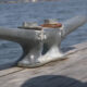 Metal mooring cleat bolted to a weathered wooden dock with water in the background.