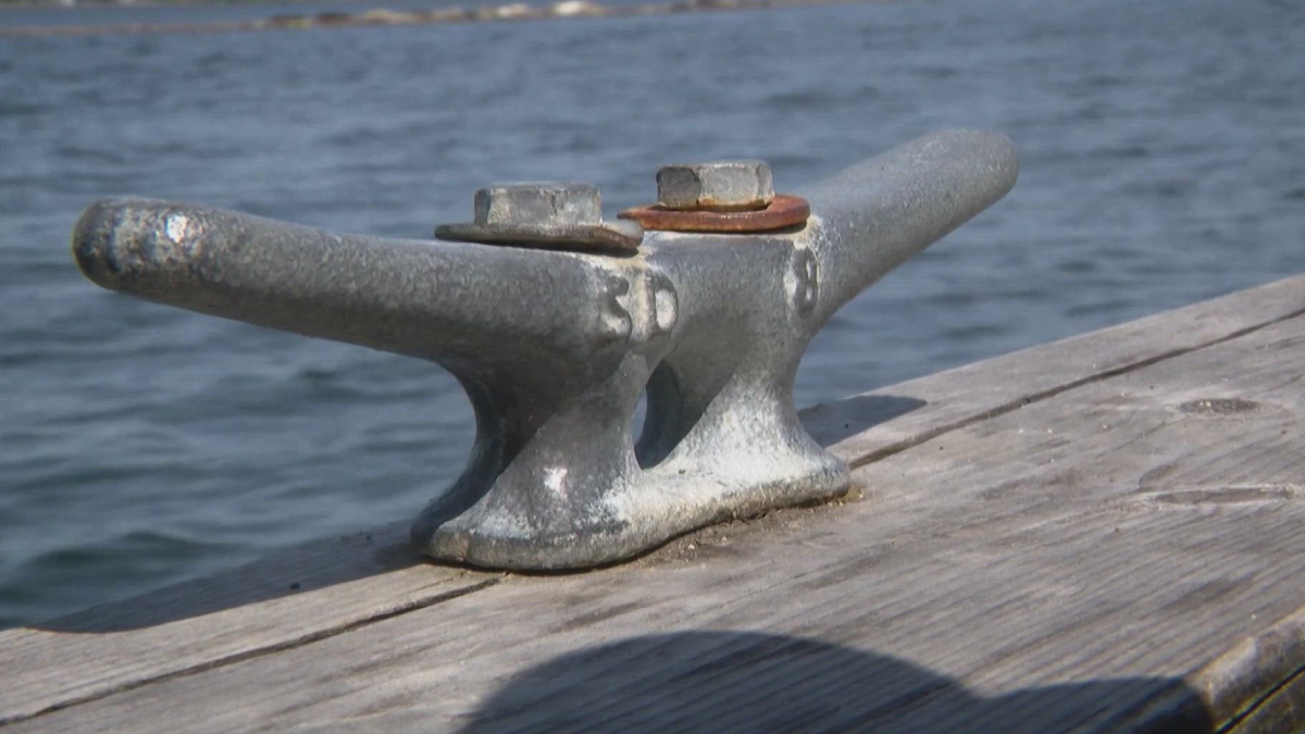 Metal mooring cleat bolted to a weathered wooden dock with water in the background.