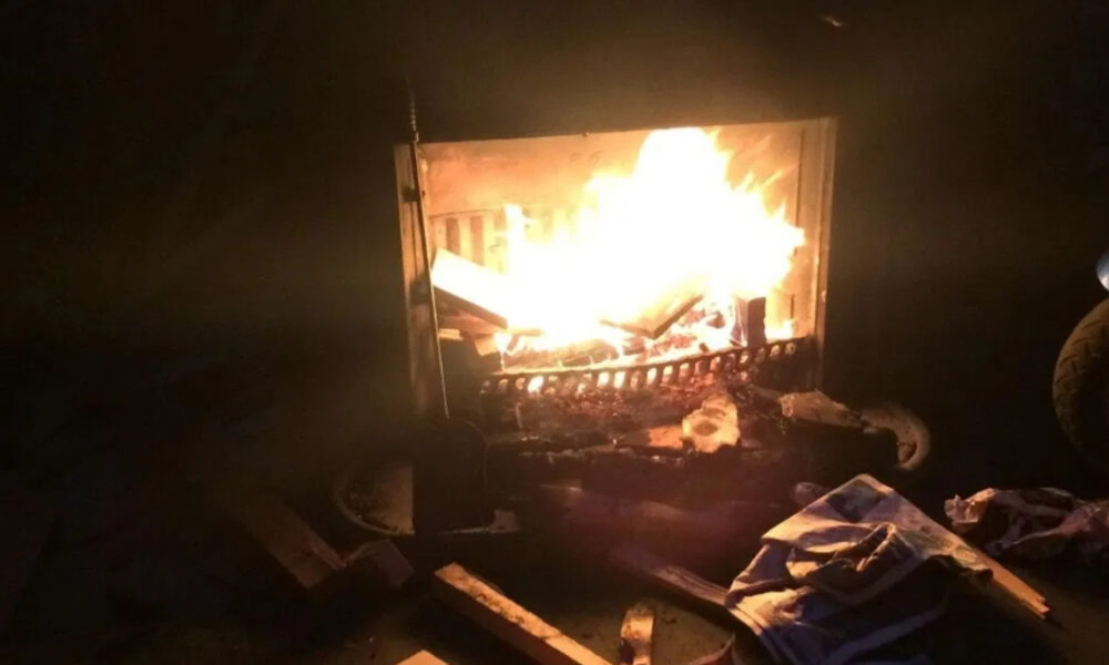 Dark room with a wood-burning fireplace; bright flames consume logs and burning embers glow in the firebox, surrounded by scattered debris on the floor.