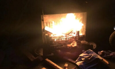 Dark room with a wood-burning fireplace; bright flames consume logs and burning embers glow in the firebox, surrounded by scattered debris on the floor.