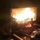 Dark room with a wood-burning fireplace; bright flames consume logs and burning embers glow in the firebox, surrounded by scattered debris on the floor.