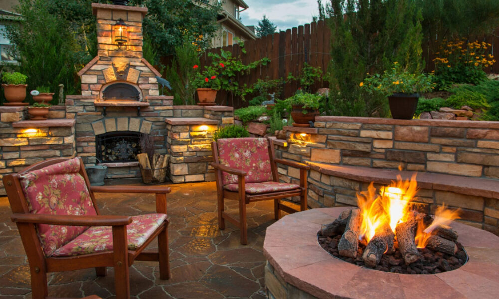 Cozy backyard with a stone outdoor fireplace, two cushioned chairs, and a glowing circular fire pit.