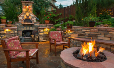 Cozy backyard with a stone outdoor fireplace, two cushioned chairs, and a glowing circular fire pit.