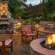 Cozy backyard with a stone outdoor fireplace, two cushioned chairs, and a glowing circular fire pit.