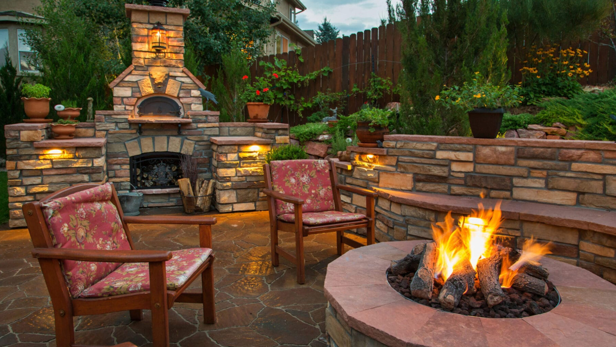 Cozy backyard with a stone outdoor fireplace, two cushioned chairs, and a glowing circular fire pit.