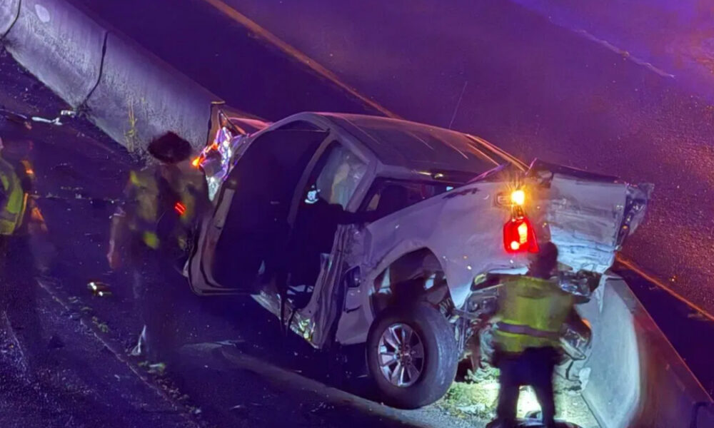 White pickup truck crashed against a guardrail on a wet road; door open and rear damage visible as responders in reflective vests work at the scene with flashing emergency lights nearby.