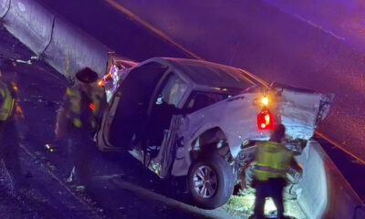 White pickup truck crashed against a guardrail on a wet road; door open and rear damage visible as responders in reflective vests work at the scene with flashing emergency lights nearby.