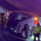 White pickup truck crashed against a guardrail on a wet road; door open and rear damage visible as responders in reflective vests work at the scene with flashing emergency lights nearby.
