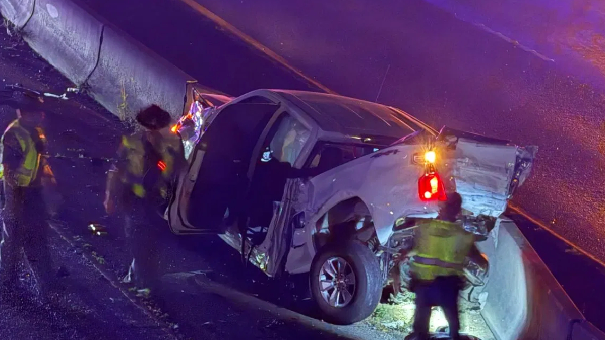 White pickup truck crashed against a guardrail on a wet road; door open and rear damage visible as responders in reflective vests work at the scene with flashing emergency lights nearby.