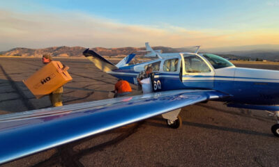 Blue single-engine plane on a sunlit runway with cargo doors open as workers load boxes at sunset.