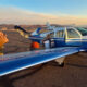 Blue single-engine plane on a sunlit runway with cargo doors open as workers load boxes at sunset.