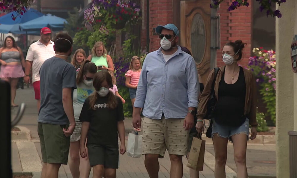 Group of people walking on a sunny street wearing face masks; shoppers carry bags on a brick-paved sidewalk.