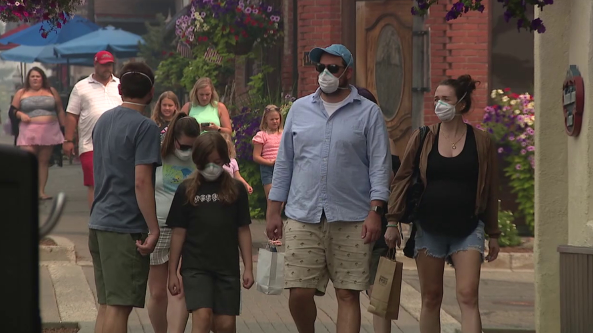 Group of people walking on a sunny street wearing face masks; shoppers carry bags on a brick-paved sidewalk.