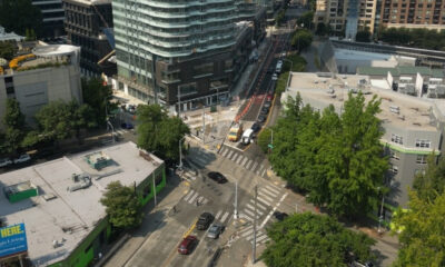 Aerial view of a busy urban intersection with cars, buses, and tall modern buildings surrounded by trees.