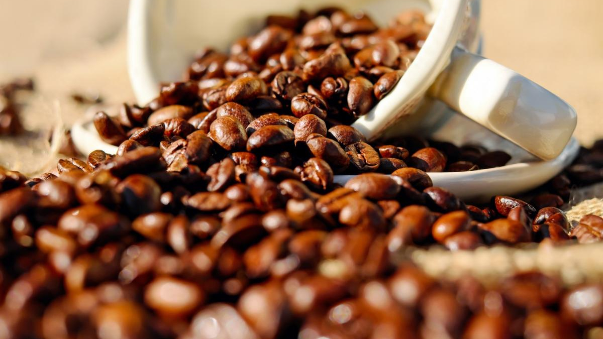 Brown roasted coffee beans spilling from a white ceramic cup onto a rustic surface.