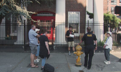 Group of people standing on a sidewalk and listening to a man with papers as a police officer in a black shirt talks nearby, with a yellow fire hydrant in the foreground.