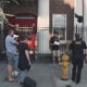 Group of people standing on a sidewalk and listening to a man with papers as a police officer in a black shirt talks nearby, with a yellow fire hydrant in the foreground.