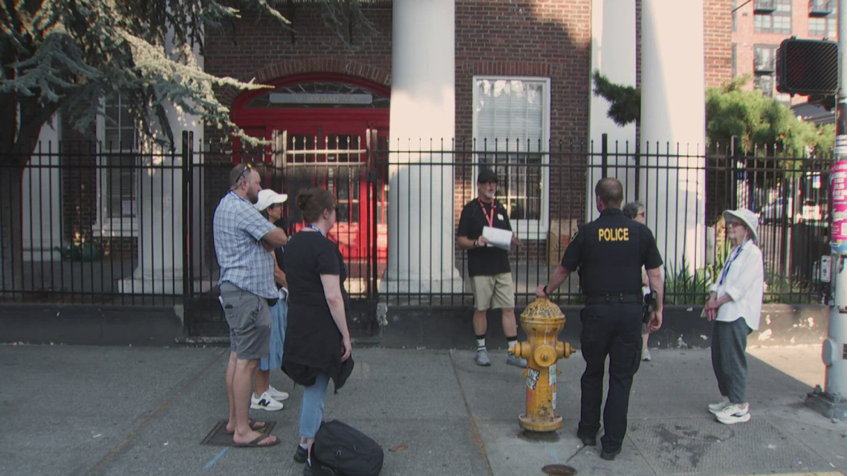 Group of people standing on a sidewalk and listening to a man with papers as a police officer in a black shirt talks nearby, with a yellow fire hydrant in the foreground.