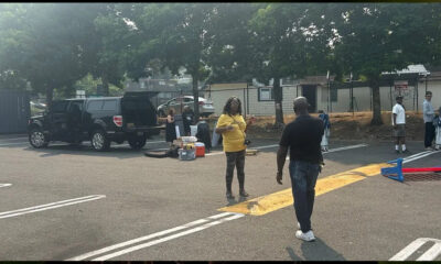 People set up gear in a parking lot beside a black SUV with open doors; a woman in yellow speaks to a man approaching.