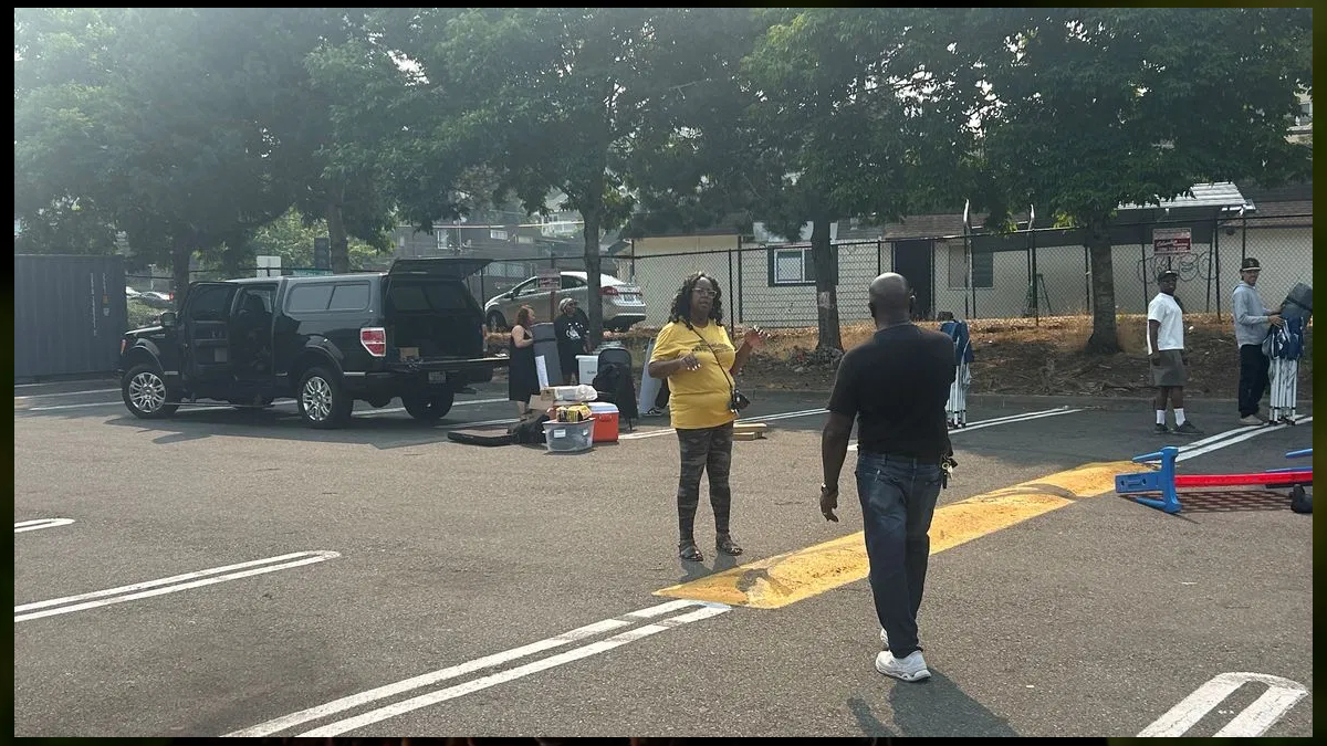 People set up gear in a parking lot beside a black SUV with open doors; a woman in yellow speaks to a man approaching.
