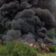 Thick black smoke rising from a grass and brush fire in a wooded area with trees in the background