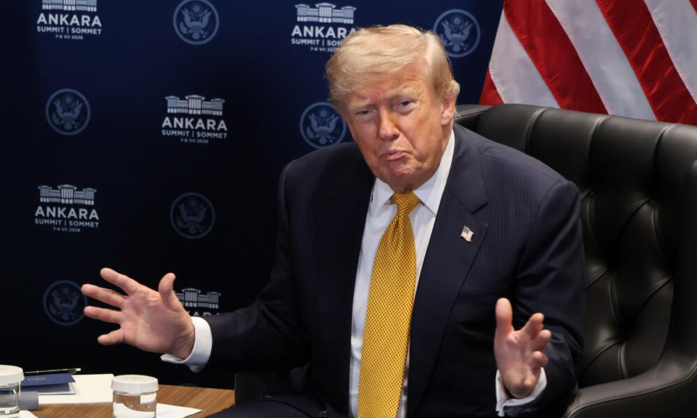 A suited man with a yellow tie sits at a table, gesturing with his hands during a briefing, with a US flag in the background.