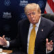 A suited man with a yellow tie sits at a table, gesturing with his hands during a briefing, with a US flag in the background.
