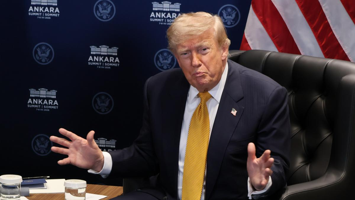 A suited man with a yellow tie sits at a table, gesturing with his hands during a briefing, with a US flag in the background.