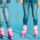 Two people wearing ripped jeans in pink inline skates stand on a bright blue backdrop, holding hands.