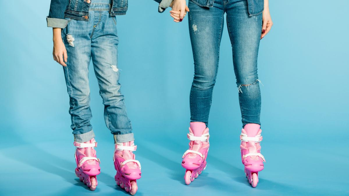 Two people wearing ripped jeans in pink inline skates stand on a bright blue backdrop, holding hands.