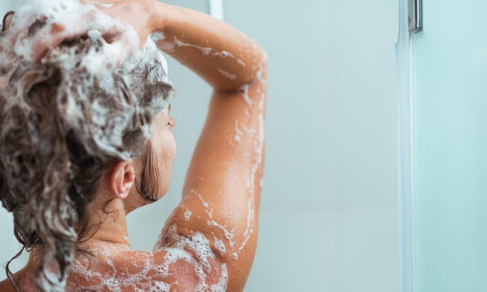 Person washing their hair in the shower, with white shampoo foam on the head and shoulders (back view)