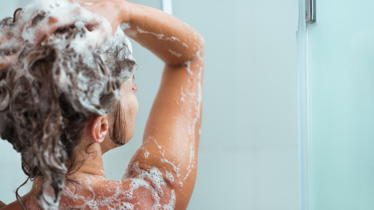 Person washing their hair in the shower, with white shampoo foam on the head and shoulders (back view)