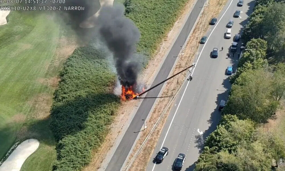 Car on fire in the median of a divided highway, with thick black smoke rising over the scene and nearby traffic passing by (in aerial view).