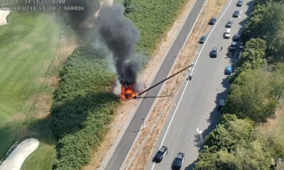 Car on fire in the median of a divided highway, with thick black smoke rising over the scene and nearby traffic passing by (in aerial view).