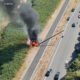 Car on fire in the median of a divided highway, with thick black smoke rising over the scene and nearby traffic passing by (in aerial view).