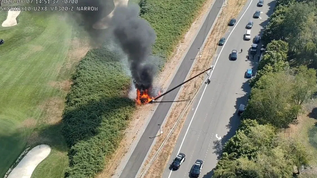 Car on fire in the median of a divided highway, with thick black smoke rising over the scene and nearby traffic passing by (in aerial view).