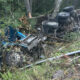 Blue semi-truck overturned on a forested hillside, with wheels and undercarriage visible among brush and debris (accident scene).